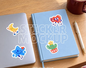 May include: A light blue notebook and a silver laptop featuring colourful cartoon stickers of a yellow crescent moon, a red heart, a blue star, and a green cactus. A white pen and a dark red mug are placed nearby on a wooden surface.