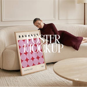 May include: A framed poster with a Bauhaus design featuring pink circles on a red background. The poster is in a light wood frame and has the words "BAUHAUS", "AUSSTELLUNG", and "1923" printed on it. The poster is leaning against a cream-colored sofa.
