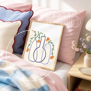 Op de afbeelding: Een ingelijste kunstdruk met een minimalistische illustratie van vazen, oranje bloemen en groene wijnranken. Het kunstwerk ligt op een bed met roze en blauwe kussens, een blauw-wit geblokte deken en een roze sprei. Het woord "ooto" staat rechtsboven.