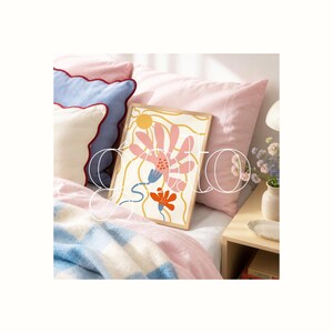 Op de afbeelding: Een ingelijste kunstprint met een bloemmotief in roze, oranje en blauwe tinten. Het kunstwerk heeft een cr&egrave;mekleurige achtergrond en een houten lijst. De print wordt gepresenteerd op een bed met roze kussens en een blauw-witte deken.