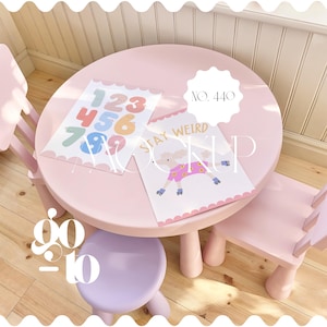 May include: A pink children's table and chairs set. Two illustrated prints are on the table, one with numbers and the other with a cartoon character and the words "Stay Weird". A small purple stool is in front of the table.