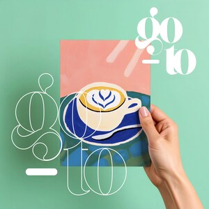 Op de afbeelding: Een hand die een print vasthoudt met een gestileerde koffiekop met latte art. De kop is wit met een blauwe schotel, tegen een roze en blauwe achtergrond. De woorden "go to" zijn in het wit overgelegd.