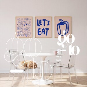 Peut inclure: Trois impressions d'art encadr&eacute;es avec un fond rose clair et des illustrations bleues sont accroch&eacute;es au-dessus d'une table blanche et de chaises transparentes. Une impression dit "LET'S EAT" en lettres bleues. Un chien se repose sur une chaise.