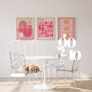 Peut inclure: Trois affiches encadr&eacute;es aux tons roses et dor&eacute;s sont accroch&eacute;es au-dessus d'une table blanche et de chaises transparentes. Les affiches pr&eacute;sentent les mots "BAUHAUS" et "GOOD TIMES". Un petit chien se repose sur une des chaises.