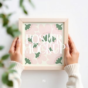 May include: A square, light wood frame displays a poster with a floral design. The poster has pink flowers and green leaf shapes on a cream background. The words "POSTER MOCKUP" are printed in white. The frame is held by hands wearing a white sweater.