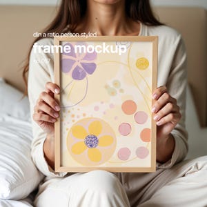 Puede incluir: Una persona sostiene un marco de madera con una impresión abstracta colorida. La impresión presenta flores amarillas, rosas y moradas y círculos sobre un fondo amarillo claro. El marco está etiquetado como "frame mockup no. 057".