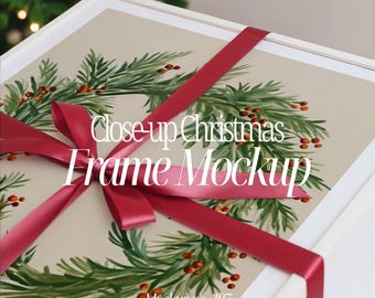 Op de afbeelding: Een wit omlijst kerstthema mockup met een bordeauxrood lint in een strik geknoopt. Het kunstwerk toont een groene krans met rode bessen. Tekst op het frame luidt "Close-up Christmas Frame Mockup" en "Mockup no. 487".