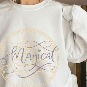 May include: White sweatshirt with the word "Magical" in a cursive font, overlaid on a gold geometric design. The word "WHITE" is in a black circle at the bottom. The sweatshirt has a crew neck.