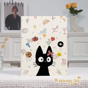 May include: A cream-colored notebook featuring a black cat illustration with a pink bow. The cover is decorated with various whimsical drawings, including a strawberry, stars, and a cookie. The notebook is a unique and artistic item.