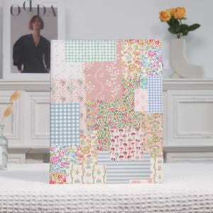 May include: A hardcover journal with a patchwork design. The cover features a collage of floral, gingham, and striped patterns in pastel pink, blue, and green. The journal is displayed on a white surface.