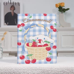 May include: A notebook with a blue and white gingham pattern featuring a cartoon character holding a basket of cherries. The word "Cinnamoroll" is written in a cursive font above the character.