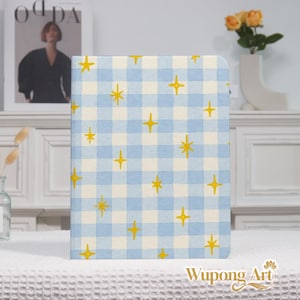 May include: A light blue and white checkered notebook cover with yellow star accents. The notebook is standing upright on a white surface. The notebook has a watercolor design and is a decorative item.