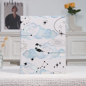 May include: A notebook with a celestial design. The cover features light blue watercolor clouds, stars, a crescent moon, and constellations on a white background. Black line art elements include planets and swirling patterns.