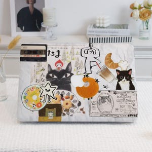 May include: A white laptop case adorned with stickers of a black cat, a cassette tape, a star, and a donut. The case features Japanese text and food illustrations.