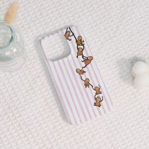 May include: A rectangular phone case featuring a light pink and white vertical striped pattern. A chain of seven brown cartoon monkeys is illustrated hanging from a vine that curves across the centre of the case, with one monkey sitting on a yellow swing.