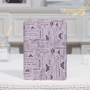 May include: A rectangular, lavender-colored notebook with a vintage French newspaper print design. The cover features script, keys, butterflies, and the text "J. GIRAUD FILS" and "LE DE PARIS". The notebook is on a white surface.