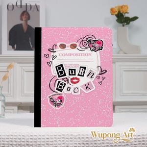May include: Pink composition notebook with a black spine. The cover features the words "BURN BOOK" in a collage style, along with lipstick, sunglasses, and hearts. The notebook has the text "COMPOSITION" and "ON WEDNESDAYS WE WEAR PINK".