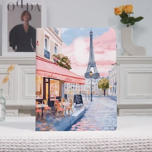 May include: A notebook featuring a Parisian cafe scene with the Eiffel Tower in the background. The cover art depicts a cafe with a red awning, outdoor seating, and a handwritten menu. The sky is pink and blue.