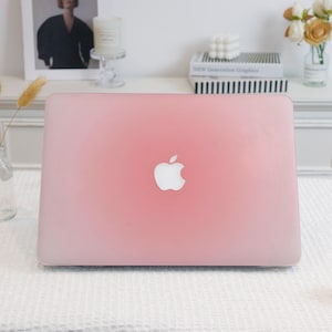 May include: A pink laptop computer with the Apple logo centered on the lid. The laptop has a gradient color, transitioning from a darker pink at the edges to a lighter pink in the center. The laptop is on a white surface.