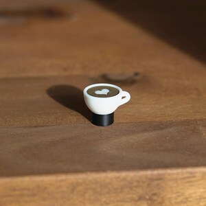 Op de afbeelding: Een kleine, witte koffiekop met een hartvormig ontwerp in de koffie. De kop staat op een zwarte basis. De koffiekop is een miniatuurversie van een latte.