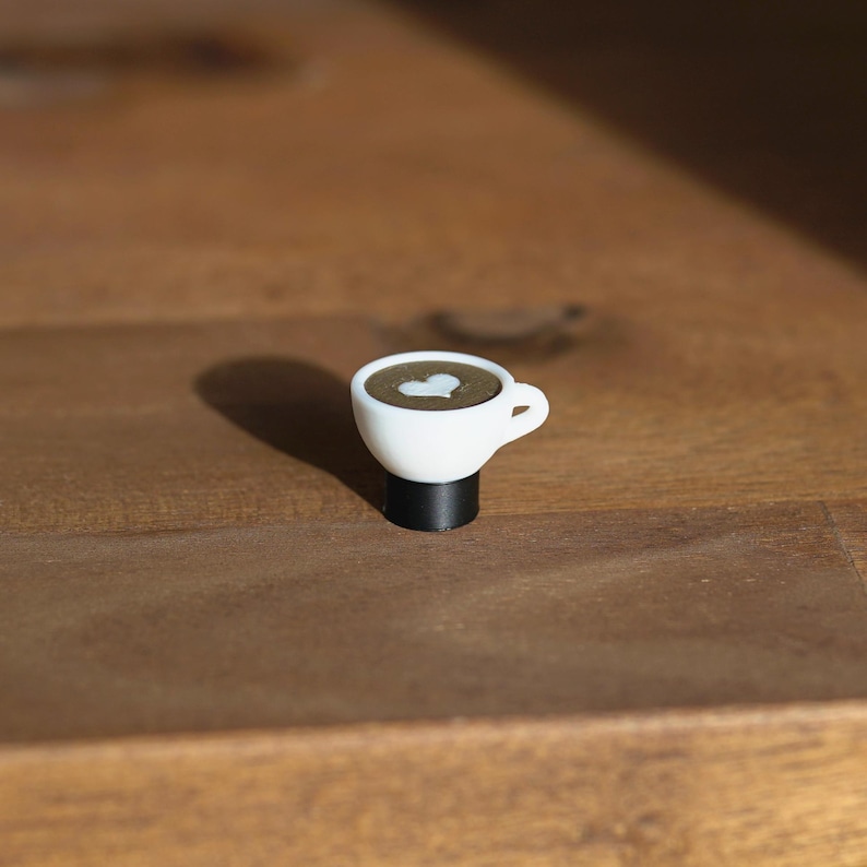 Op de afbeelding: Een kleine, witte koffiekop met een hartvormig ontwerp in de koffie. De kop staat op een zwarte basis. De koffiekop is een miniatuurversie van een latte.