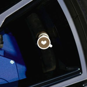 Op de afbeelding: Een witte en bruine bandventieldop met een koffiekopje ontwerp en een hartvormige schuimkunst. De dop staat tegen een donkere achtergrond, met een glimp van een blauw auto-onderdeel.