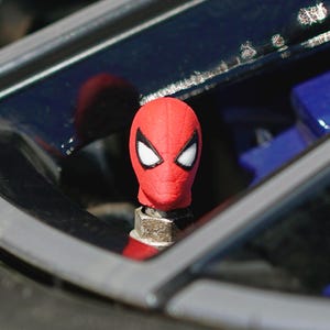 Puede incluir: Un tapón de válvula de neumático de Spider-Man rojo con detalles de ojos blancos. La tapa está montada en una válvula de metal plateado. El fondo incluye el interior y el exterior de un coche.