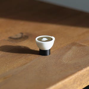 Op de afbeelding: Een kleine witte koffiekop met een zwarte basis, met een hartvormig ontwerp in de koffie. De kop staat op een houten oppervlak, wat een warme uitstraling cre&euml;ert. Het miniatuurformaat suggereert een decoratief of nieuwigheid item.