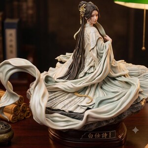 May include: A detailed figurine of a person with long dark hair styled in an updo with gold ornaments, wearing a flowing pale green robe with intricate silver embroidery. The figure sits on a dark base with the Chinese text 白仕詩 inscribed on a plaque.