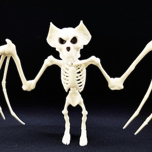 May include: A plastic, off-white bat skeleton figurine with outstretched wings. The figurine has a detailed rib cage, bat-like ears, and dark eye sockets. The Halloween decoration is set against a black background.