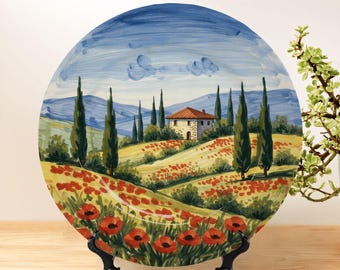 May include: A round ceramic decorative plate featuring a hand-painted landscape of a stone house with a red roof, surrounded by rolling hills, tall cypress trees, and a field of vibrant red poppies under a blue sky with soft white clouds.