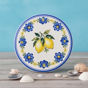 May include: A round ceramic plate with a white background featuring a central illustration of two yellow lemons with green leaves. A decorative border of blue and yellow flowers encircles the lemons, framed by thin blue circular lines.
