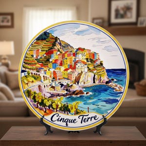 Puede incluir: Un plato decorativo de cerámica con una colorida escena de un pueblo costero con el texto "Cinque Terre". El plato tiene un borde amarillo y se exhibe en un soporte negro. La obra de arte representa edificios en un acantilado con vistas al mar.