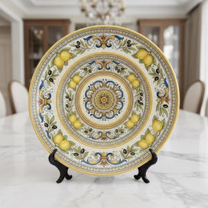 May include: A decorative ceramic plate with a lemon and olive design. The plate features a yellow, blue, and white colour scheme with a detailed pattern of lemons, olives, and floral elements. The plate is displayed on a black stand.