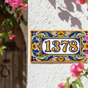May include: Rectangular ceramic address plaque with the number 1378 in yellow. The plaque has a yellow border and is decorated with blue, green, and yellow floral patterns. The plaque is mounted on a white textured wall.