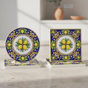 Może przedstawiać: Dwa ceramiczne podstawki, jedna okrągła i jedna kwadratowa, z motywem cytryny. Podstawki mają białą podstawę z niebiesko-ż&oacute;łtym wzorem i złotą obw&oacute;dką. Wz&oacute;r cytryny jest wyśrodkowany na białym tle.