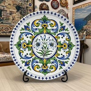 May include: A decorative ceramic plate with a floral design. The plate features a white background with a central medallion of white lilies and blue and green foliage. The border has a repeating pattern of blue and yellow accents.