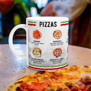 May include: White ceramic mug with a colorful illustration of four different pizzas. The mug has the word "PIZZAS" printed in black at the top. The pizza names are listed below each pizza image, along with a list of ingredients. The mug is sitting on a table with a pizza in the foreground.