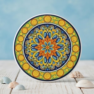 May include: A circular ceramic coaster with a colourful floral pattern. The coaster has a central flower in orange, yellow, and blue, set against a navy blue background, with a green border and yellow spiral details. It rests on a white wooden surface.