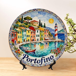 May include: A decorative ceramic plate depicting the Italian coastal town of Portofino. The plate shows colorful buildings, boats, a lighthouse, and the word "Portofino". The plate is displayed on a black stand and has a detailed border.
