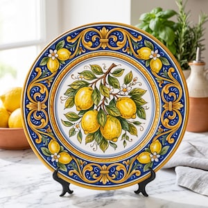 May include: A round ceramic decorative plate featuring a central illustration of a lemon branch with four yellow lemons, green leaves, and white blossoms. The border displays a blue and yellow scrollwork pattern with additional lemon motifs.
