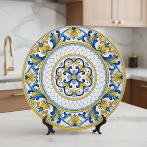 Puede incluir: Un plato decorativo de cerámica con un diseño vibrante. El plato presenta un motivo floral central en azul y amarillo, rodeado de patrones azules y amarillos ornamentados sobre un fondo blanco. El plato se exhibe sobre un soporte negro.