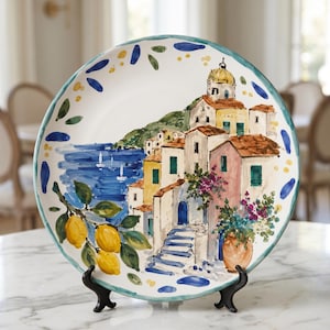 Puede incluir: Un plato redondo de cerámica con una escena costera pintada a mano que muestra casas coloridas de estilo mediterráneo, un mar azul con veleros y una rama de limones amarillos. El borde está decorado con pinceladas azules y amarillas.