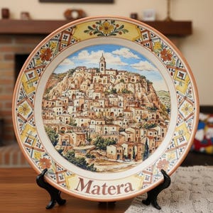 Puede incluir: Un plato decorativo redondo de cerámica con una escena pintada a mano de la ciudad de Matera, Italia. El centro representa una ciudad en la ladera con edificios de piedra y un campanario. El borde tiene motivos florales amarillos, naranjas y verdes. La palabra Matera está impresa en la parte inferior.