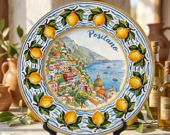 May include: A round ceramic decorative plate featuring a hand-painted coastal scene of Positano, Italy, with colorful buildings and blue water. The border is decorated with a repeating pattern of yellow lemons and green leaves on a blue wavy background.
