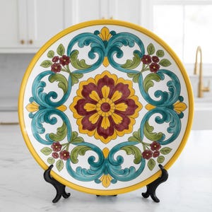 May include: A decorative ceramic plate with a yellow rim and detailed floral design. The plate features a central flower in burgundy and yellow, surrounded by teal and green scrollwork and small red flower clusters. The plate is displayed on a black stand.