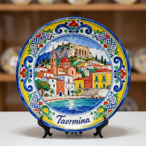 May include: A decorative ceramic plate featuring a colorful depiction of Taormina, Italy. The plate has a yellow border with blue and red floral designs. The central image shows buildings, a church, and the sea. The plate is displayed on a black stand.