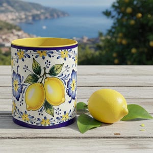 May include: A ceramic container with a yellow interior, decorated with lemons and blue and yellow floral patterns. A fresh lemon and green leaves sit on a wooden surface next to the container. The background shows a coastal landscape.