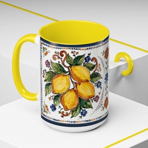 May include: A ceramic coffee mug with a yellow interior and handle. The exterior features a white background with a painted design of lemons, green leaves, and colorful flowers. A navy blue band accents the top and bottom.
