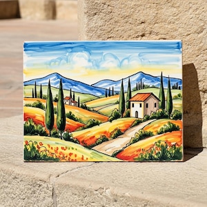 May include: A rectangular ceramic tile featuring a hand-painted landscape scene. The artwork depicts rolling hills, cypress trees, and houses under a blue sky with clouds. The color palette includes shades of yellow, orange, green, and blue.
